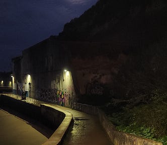 A walkway next to a hill with a low wall. The full moon shines overhead and a bit of the Tagus river can be seen to one side.