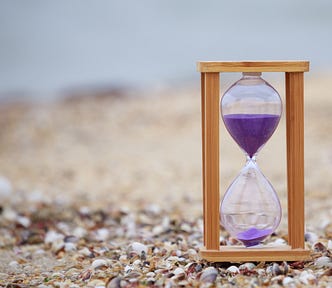 Close-up photo of a wooden hourglass with purple sand in it. Background is of a beach.