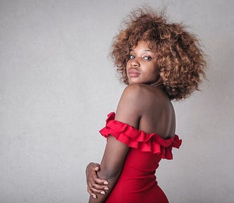 A young black woman with brown, curly hair, wearing a red off-the-shoulder dress. She is looking over her shoulder at the camera.