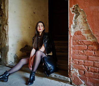 A young woman sitting in a doorway in a rundown alley. She has straight, black hair, and red lipstick. She is wearing a black leather jacket, sheer black tights, and black boots.