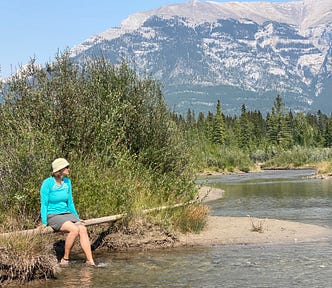 The author sits on a small bridge with her feet in the water of a river with mountains in the background.