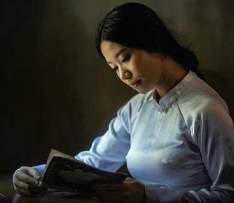 Sepia tinged photo of a young Asian woman in a high-necked white silk blouse reading a book open before her.