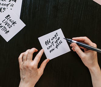 A pair of white hands with a calligraphy pen. They have written Old ways won’t open new doors. In the background is a note they have written which says Black lives matter.