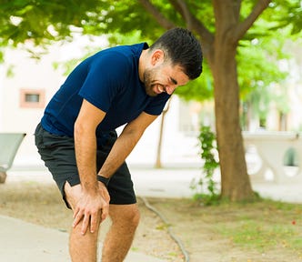 Man in running clothes grimacing in pain, clutching his knee outside in a park