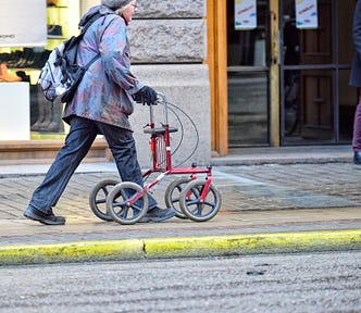 An older woman walking with the support of a rollator on the sidewalk.