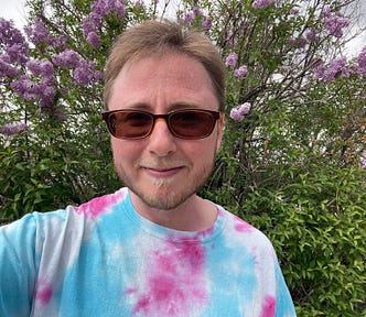 The author is wearing trans colors and standing in front of a flowery bush. He’s wearing sunglasses and has short brown hair with a mustache and short beard. He’s smiling slightly.