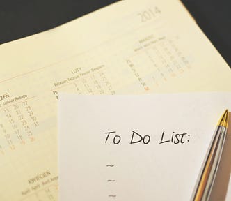 In the foreground: a “To Do List” with three empty bullet points, sitting beneath a silver and gold pen. In the background: A planner with calendars written out across it.