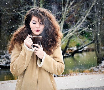 Girl, long brunette hair, winter coat, background woods and a river, blows steam off a cup of coffee