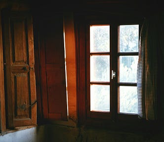 Morning light slipping through a slightly fogged wooden window, the glass streaked and soft at the edges, the room dim and quiet around it.