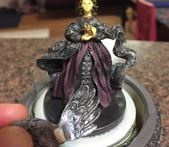 Author’s photo of a broken silver snow globe sitting on a brown countertop, featuring a light-skinned angel figure covered in silver and purple robes, holding a star. Author’s finger is shown, as she holds her broken wings in front.