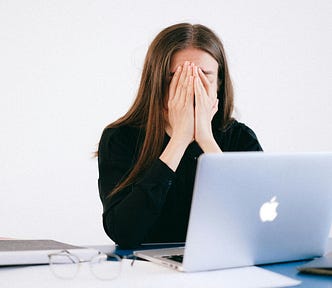 Dark-haired white woman sits in front of Macbook, eyeglasses on the desk, hands pressed over her face, looking stressed or exhausted