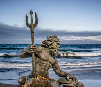 Photo of King Neptune statue on the beach with waves behind him and the shore off in a distance.
