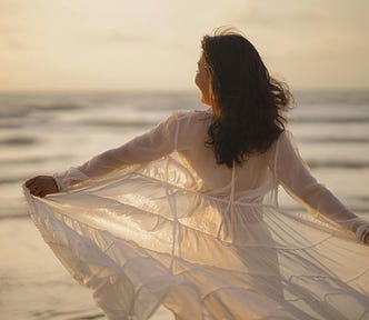 A woman standing by the sea at sunset, her dress flowing in the wind — symbolising freedom and self-liberation.