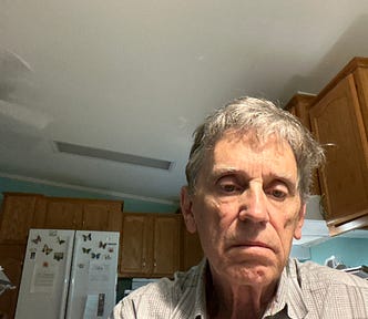 A penive older man with gray hair wearing a checked shirt is sitting in a kitchen with wooden cabinets and a white refrigerator in the background.
