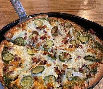 A pan of pizza with chopped dill pickles, bacon and onions.