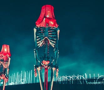 Two standard lamps made out of human skeletons with light bulbs covered by red lampshades in place of their skulls. In the background are a multitude of large strip lights, both straight and arched.