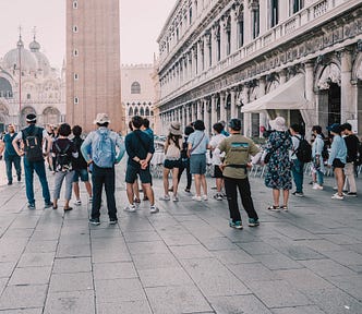 A large group of tourists, back to the camera, listening to their tour guide in St. Mark’s Square in Venice