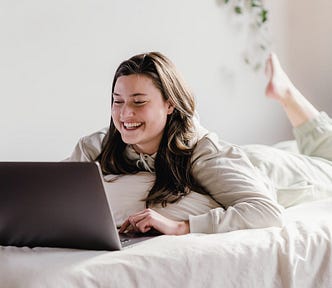 Woman laying on a bed, working on her laptop. She has one foot in the air. She is smiling. Bed and background are white and she is wearing white. Very minimalistic photograph.