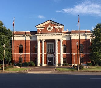 Red brick courthouse with two-story white pillars sits in the center of a typical small town square.