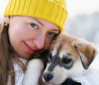 Woman with long brown hair and yellow beanie, white jacket, is smiling and hugging a husky puppy dog to her face. There is snow in the background.