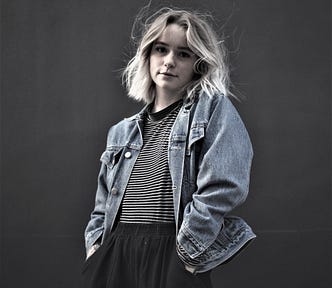 pensive girl with blonde hair wearing denim jacket and striped shirt against dark background