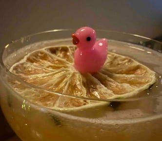 A light yellow cocktail in a clear glass topped with a dried citrus wheel, with a small pink plastic duck figurine sitting on the citrus slice.