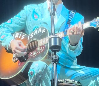 Chris Isaak, sitting on a stool playing guitar, at his concert in Australia.