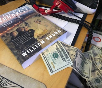A copy of a paperback book, The Accidental Terrorist by William Shunn, lies on a cluttered desk with twenty-dollar bills spilling out of it.