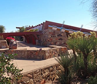 Desert brush in the foreground with a low rock wall between the garden and a large beige concrete pad. There is a low rocks ledge in a square shape with a large rock in the corner, a saguaro cactus is in the background beside a stone faced one-level sloped-roof building with a red metal portico