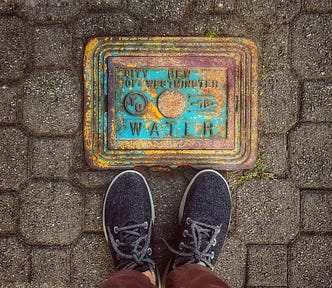 my shoes on a stone sidewalk near a patinaed water marker