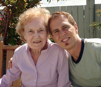 Elderly woman looks into the camera — young man has his arm around her smiling.