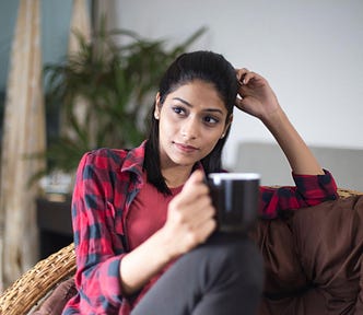 Thoughtful young woman relaxing on bean bag and drinking coffee at home