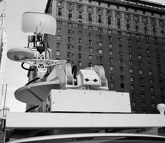 Black and white photo of an unknown contraption of technology, flanked by a skyrise building.