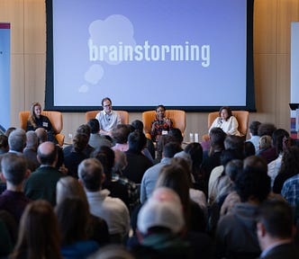 Five panelists speaking to a crowded room with a projector in the background that says “brainstorming.”