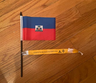 A small Haitian flag on a black stick. The flag is blue on top and red at the bottom with a white square in the middle. An organge wristband is below the flag.