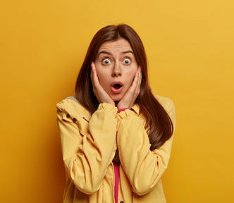 Woman with dark hair, hands clasped to her cheeks, looking surprised. Her eyes are wide and her mouth is open. She is wearing a yellow jacket, against a yellow background