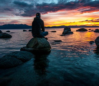 Man sitting on rock on water in front of sunset