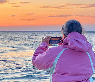 Middle aged woman wearing a pink wind breaker, on a boat, seen from behind. She is taking a picture of sunset with her phone.