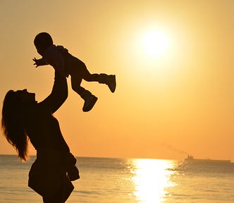 Silhouette of a woman holding a baby up over her head, while she looks up at the child, against a backdrop of a sunset over the sea