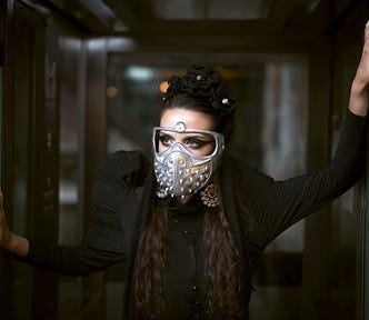 A woman wearing a dark costume and a mask is posing inside an elevator.