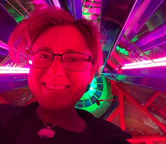 A picture is taken from below the author with a psychedelic background full of many colors simulating outer space in a creative modern art museum-like space called Meow Wolf in Denver, Colorado. The author is smiling. He’s a white man wearing glasses. His hair looks pink in the light.