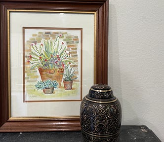 A large framed watercolor of 3 succulents and a black and gold urn sit on a gray side table