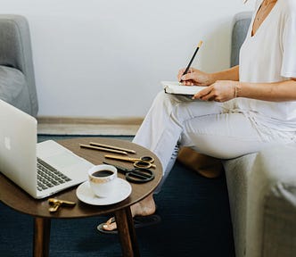 woman sits on a couch writing something in a notebook with a laptop and coffee on a small table in front of her.