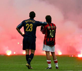 Marco Materazzi e Rui Costa observam chuva de sinalizadores no gramado do Estádio San Siro. Materazzi usa a camisa 23 azul e repousa o braço direito sobre o ombro esquerdo de Rui Costa, que veste a camisa 10 vermelho e preta.