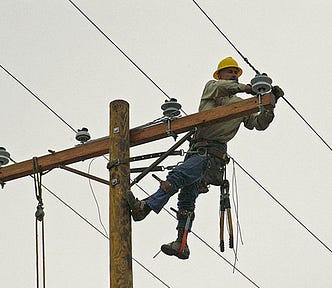 A utility worker hangs by straps from the top of a wooden utility pole, making repairs to the wires