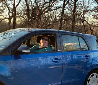 An obese white woman with short brown hair and a teal shirt sits behind the wheel of a blue and black hatchback vehicle.