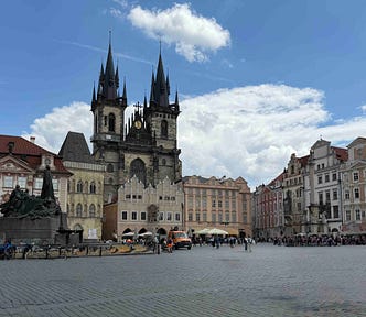 A large historic square in Prague with cobblestone paving, people walking, and outdoor seating. The Gothic Church of Our Lady before Týn rises in the background with its two tall spires, and the Jan Hus Memorial stands prominently in the center, surrounded by ornate buildings with red‑tiled roofs under a partly cloudy sky.
