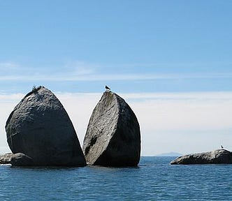 Photo of the split apple rock off the coast of ateroa/New Zealand https://en.wikipedia.org/wiki/Split_Apple_Rock The rock is large, round and grey and split in two. It is surrounded by blue water. The background has white clouds low but clear sky up above. There is a bird sittingon the right hand half of the rock.