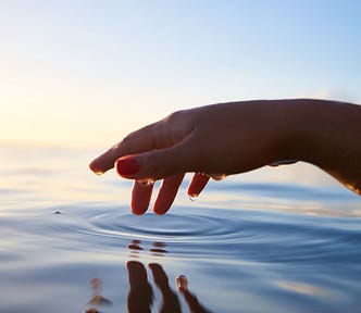 A hand, dripping wet, suspended over a glassy water surface, drops causing ripples in the surface.