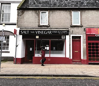 Dilapidated, 2 or 3-story stone buildings with blank windows — one houses a fish & chip shop called ‘Salt and Vinegar’, another a Chinese takeout. An indistinct person walks along the pavement in front. At the bottom of the picture, bright yellow double-lines edge the tarmac road and underline the curb.